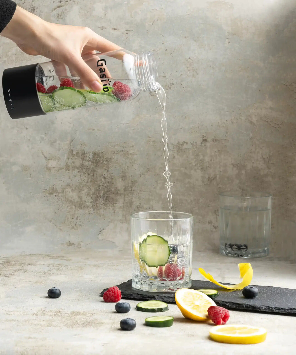 Hand pouring sparkling water from GasUp bottle into glass with cucumber, berries, and lemon slices on table.