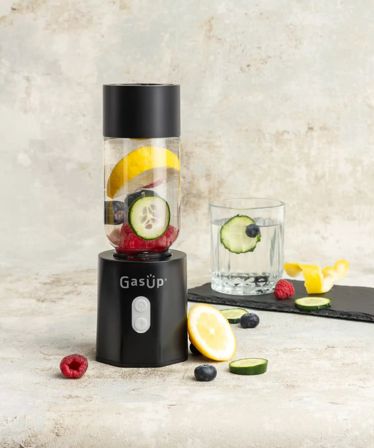 GasUp Blender Cuculo in black with fruit and cucumber slices inside, next to a glass of infused water