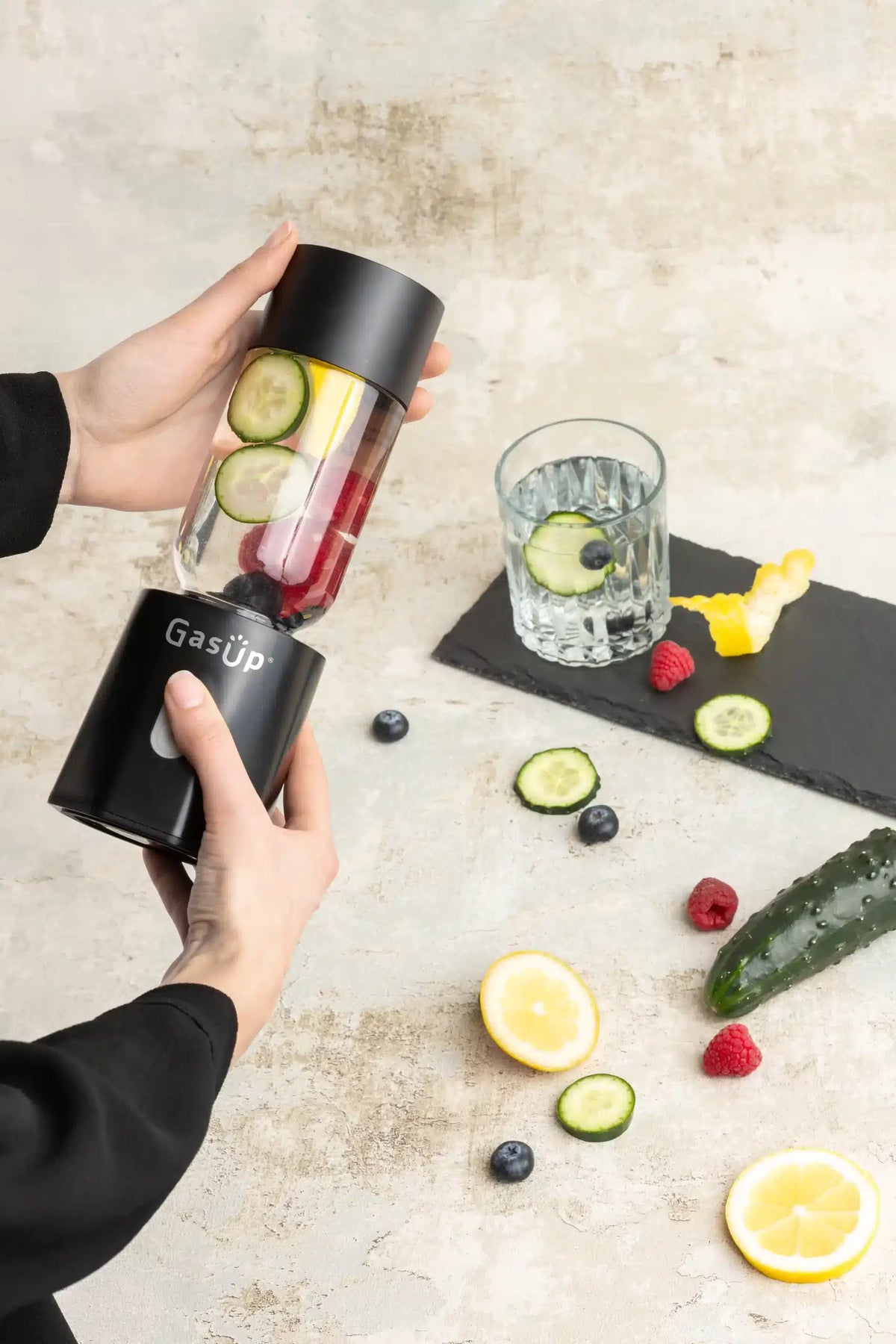 Hands holding a black GasUp Blender Cuculo blending fresh fruit and cucumber slices on a light surface.