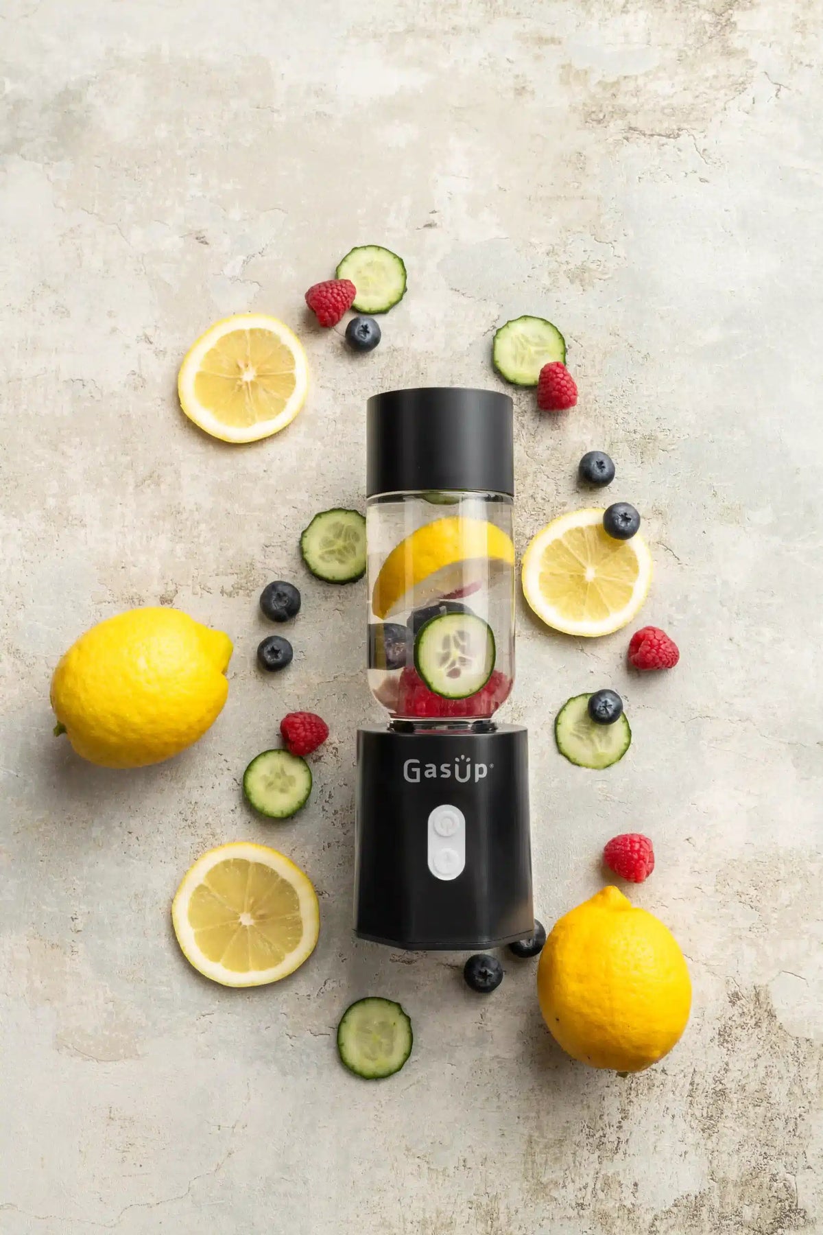 GasUp Blender Cuculo in black with lemon, cucumber, raspberry, and blueberry pieces inside and around it on a stone surface.