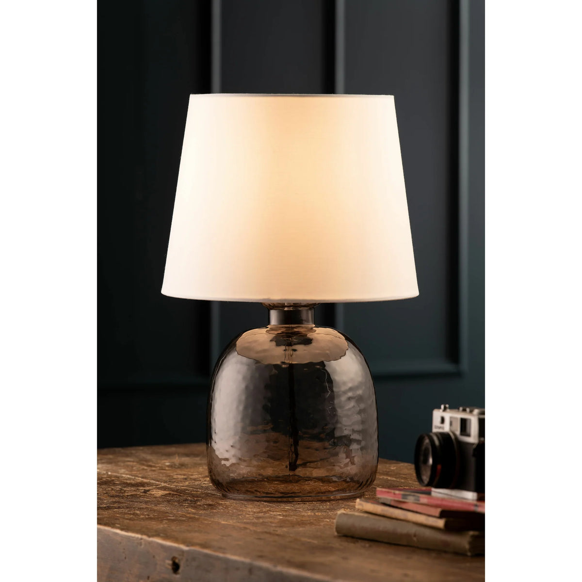 Boho table lamp with a white shade and smoky glass base on a wooden surface with books and a camera nearby.