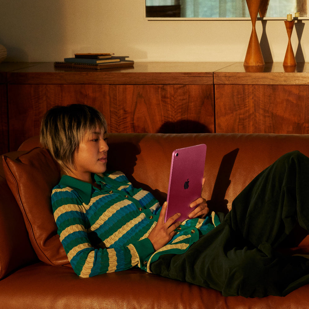 Person reclining on a brown leather couch using a purple Apple iPad A16 11" tablet.