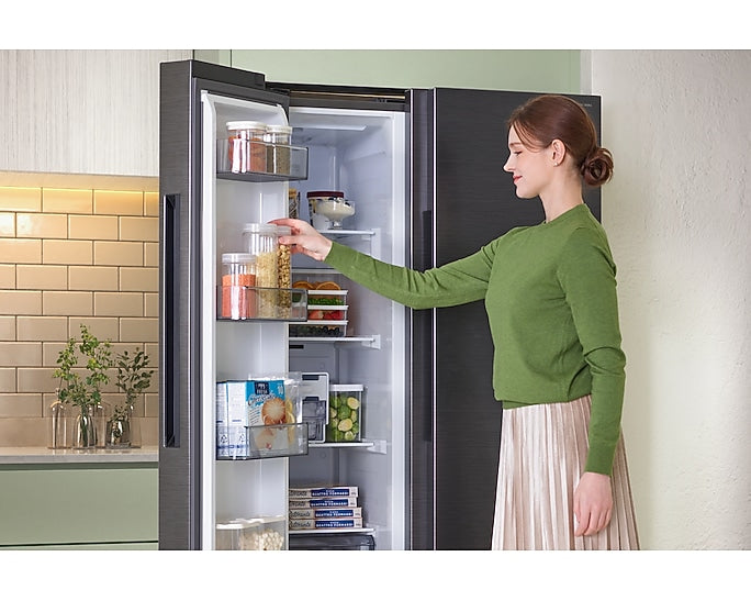 Samsung American Fridge Freezer in black steel with open door showing organized shelves and a woman reaching inside.