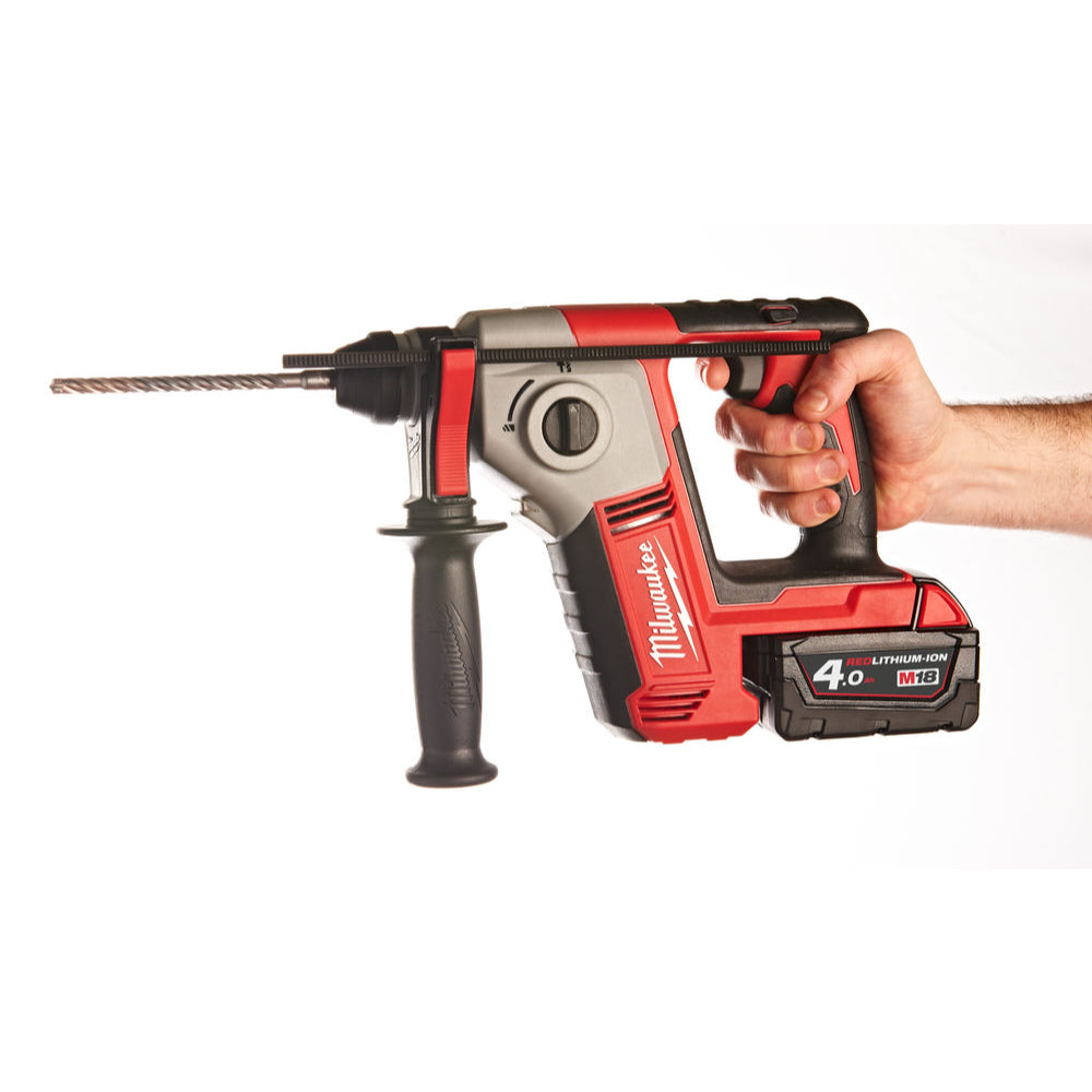 Hand holding Milwaukee M18 Compact SDS+ Hammer Drill with attached drill bit and side handle.