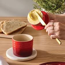 Le Creuset Cerise Butter Crock with lid lifted, showing butter inside, on wooden table with bread nearby.