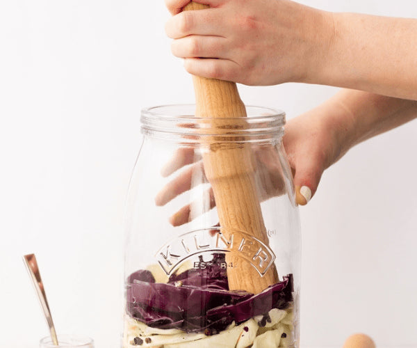 Hands using a Kilner Fermentation Packer wooden tamper inside a glass jar with cabbage and vegetables.