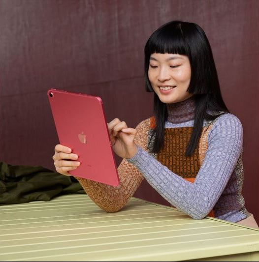 Woman holding and using a red Apple iPad 10.9" 10th Generation at a table with a maroon background.