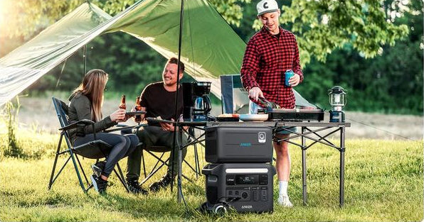 Anker BP2600 portable power expansion battery stacked on a power station at a campsite with people cooking and relaxing.