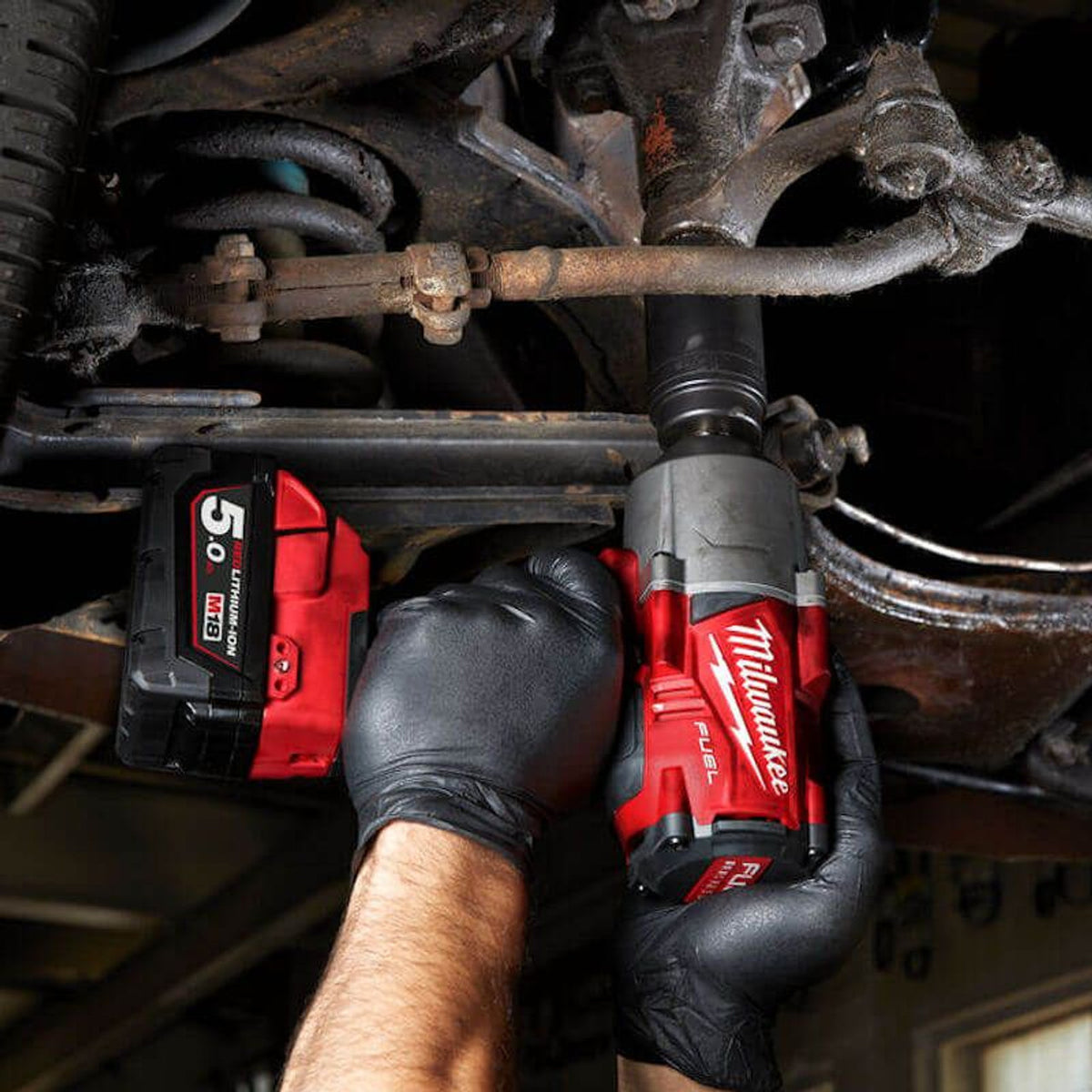 Hands wearing black gloves using a Milwaukee M18 Fuel impact wrench on a rusty vehicle part.