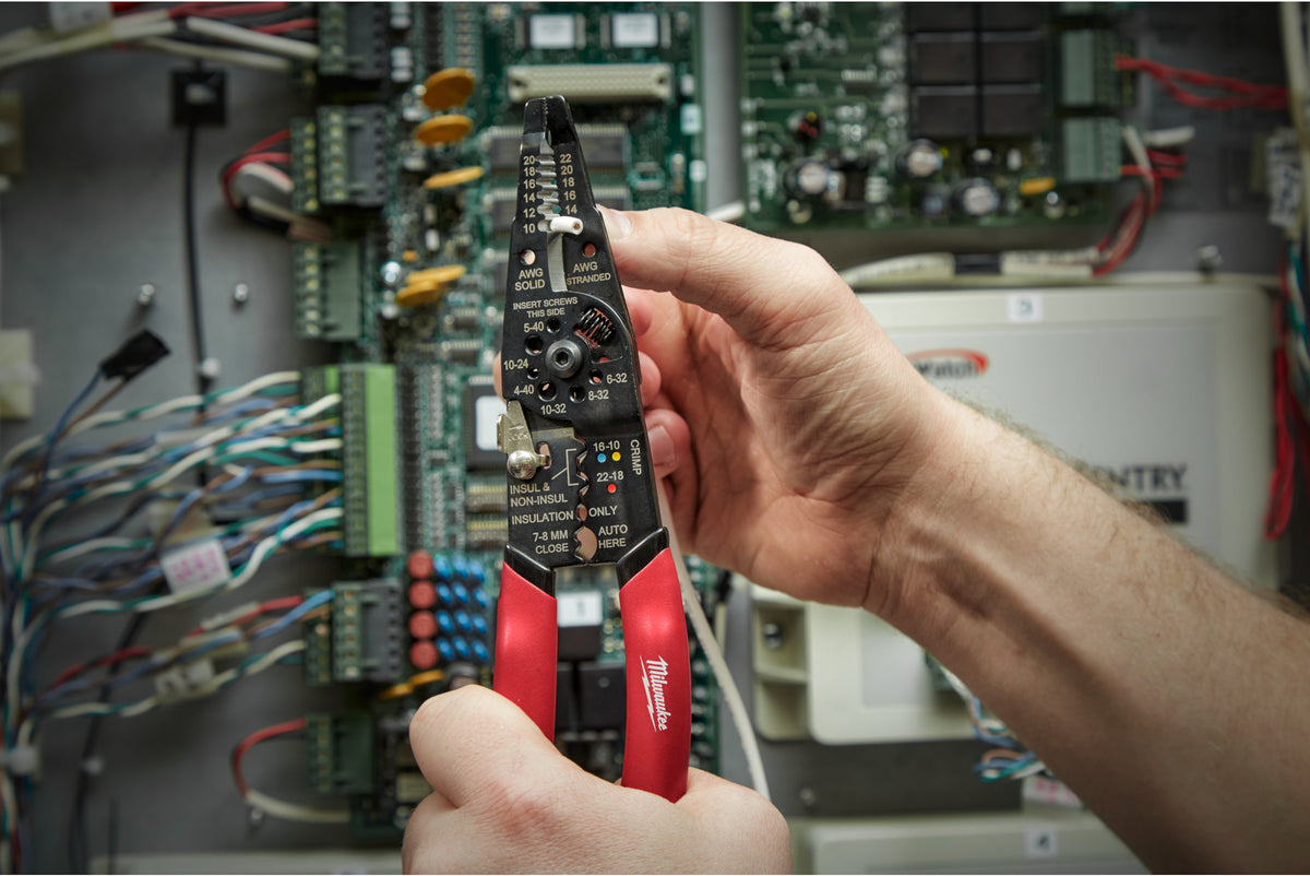 Hands holding Milwaukee Multi-Purpose Wire Stripper with Crimper stripping a white wire against a circuit board background