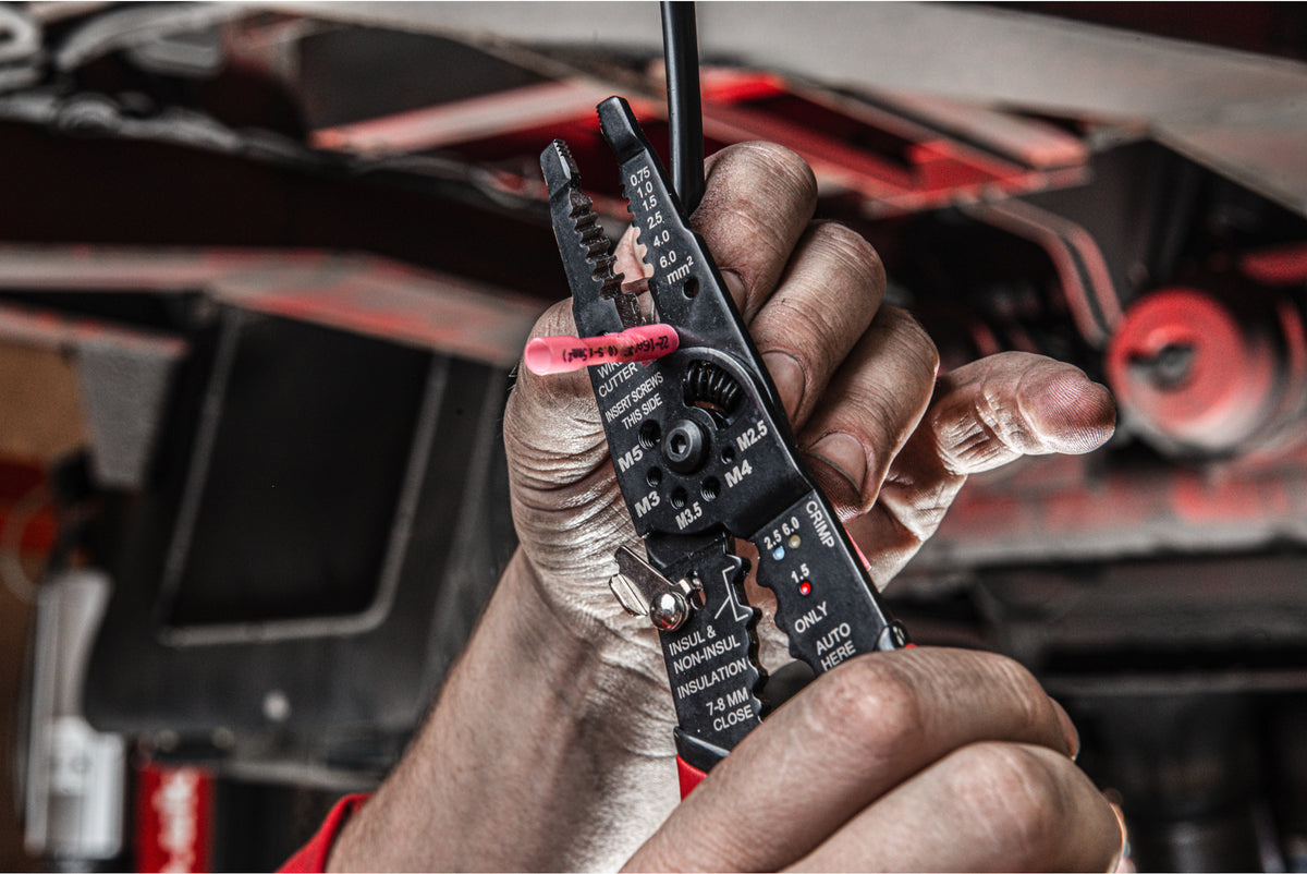 Hand holding Milwaukee multi-purpose wire stripper with crimper stripping a black wire in a workshop setting.