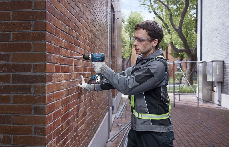 Man using Bosch 18V Combi Drill on a brick wall, wearing safety glasses and gloves outdoors