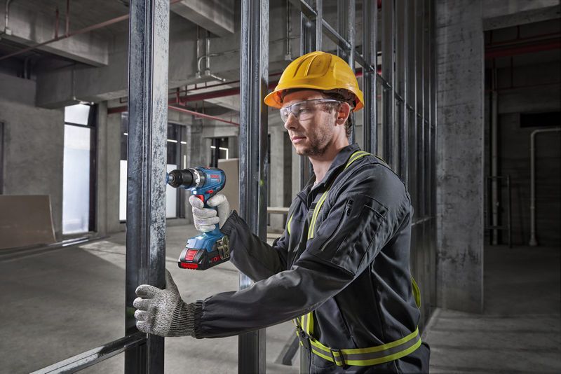 Man wearing safety gear using Bosch 18V Combi Drill on metal framing in a construction site.