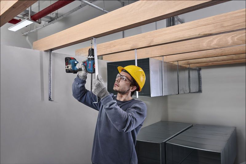 Man wearing safety gear using Bosch 18V Combi Drill to install metal brackets on wooden beams.