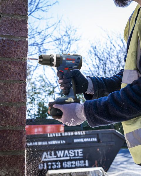Bosch 18V Combi Drill in use drilling into brick, held by a person wearing gloves and a high-visibility vest.