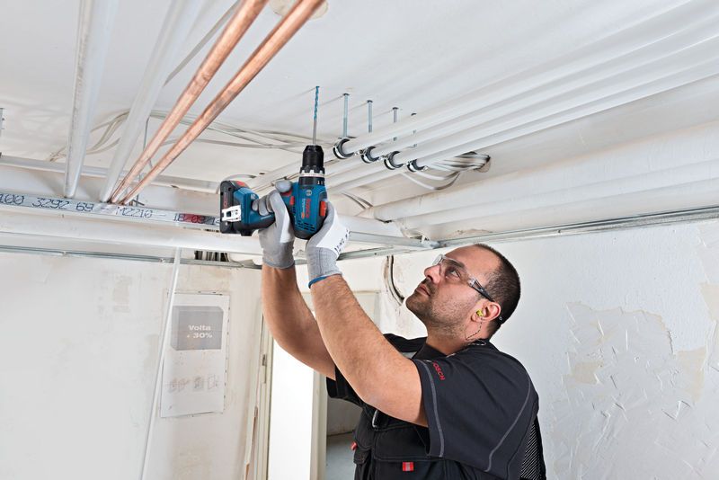 Man using Bosch 18V Combi Drill to work on pipes mounted on a ceiling in a white room.