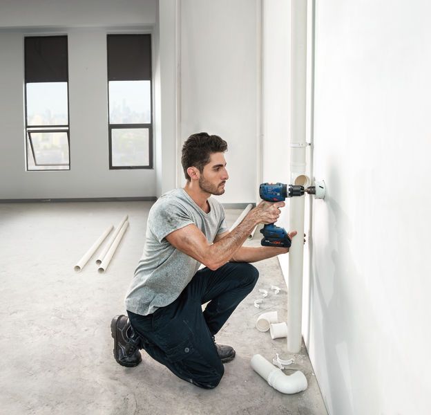 Man kneeling and using Bosch 18V Combi Drill to work on a pipe against a white wall in an empty room.