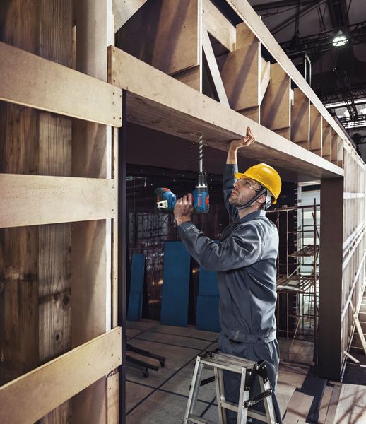Man wearing a yellow hard hat using Bosch 18V Combi Drill to drill into wooden frame on a ladder.