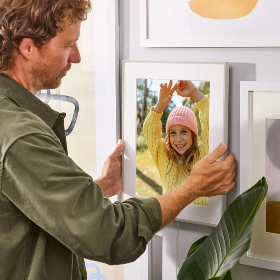 Man hanging Aura Walden 15 Inch Digital Photo Frame in clay with white paper mat on a wall displaying a smiling girl in a