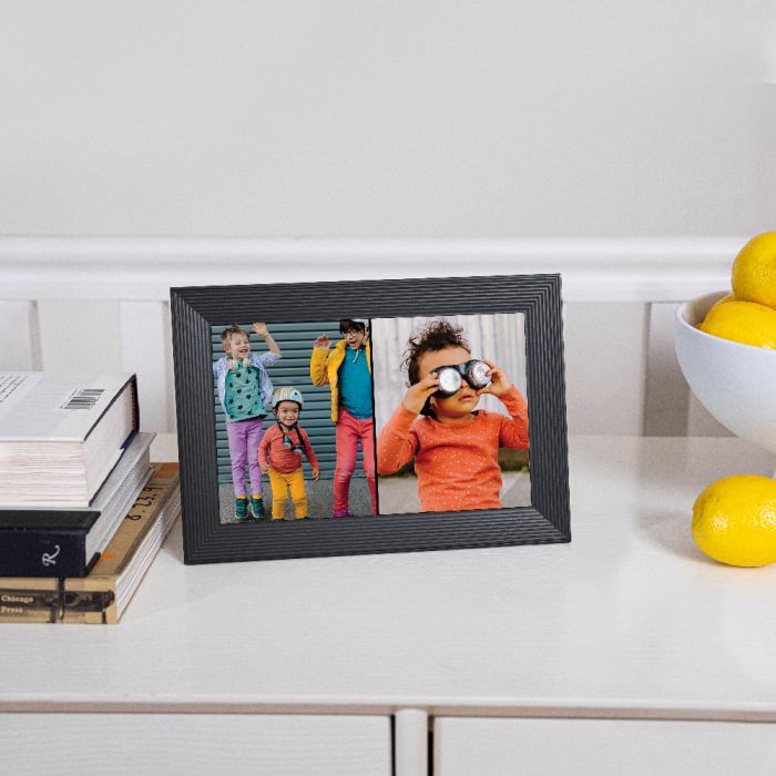 Aura Carver 10.1 inch black digital photo frame displaying two colorful photos on a white surface next to books and lemons.