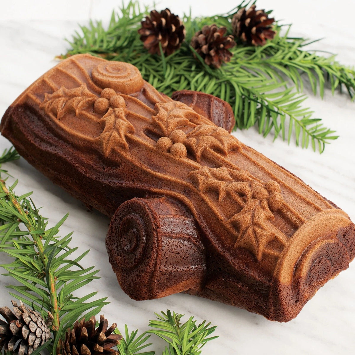 Silver Yule Log Pan with holly leaf and berry design, surrounded by pine branches and pinecones on marble surface.