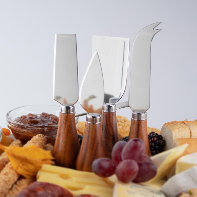 Four acacia-handled cheese knives standing upright on a cheese board with grapes, bread, and chutney.