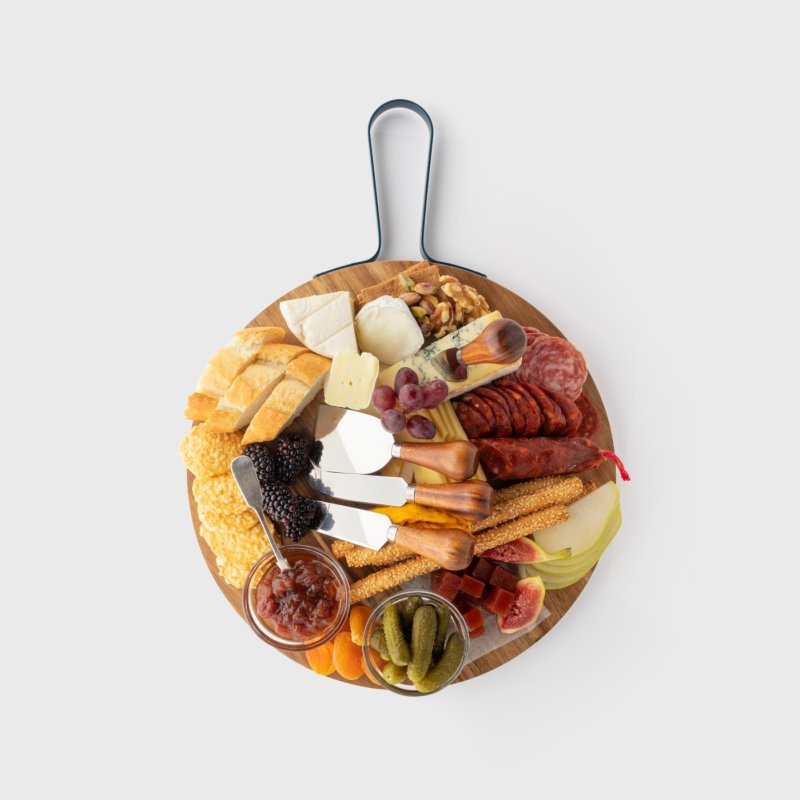 Round acacia wood charcuterie board with four cheese knives and assorted cheeses, meats, fruits, and snacks.