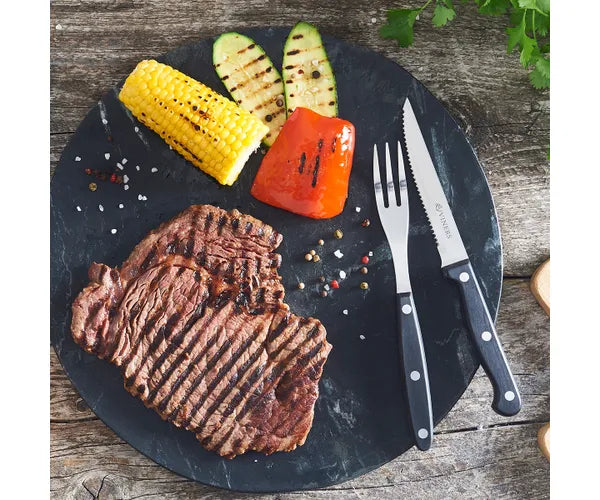 Viners 12 Piece Steak Knife & Fork Set with black handles beside grilled steak and vegetables on a black plate