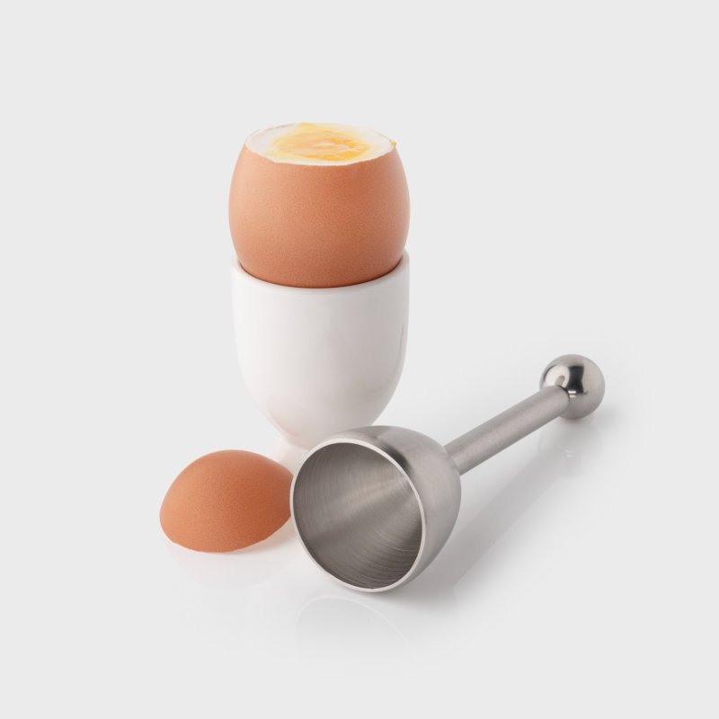 Stainless steel egg topper beside a soft-boiled egg in a white egg cup with removed eggshell top.