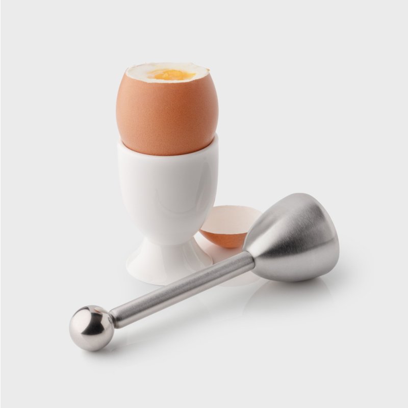 Stainless steel egg topper beside a soft-boiled egg in a white egg cup with cracked shell pieces.