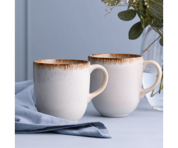 Two Mason Cash Reactive Cream Mugs with brown rim detail, placed on a light blue cloth and surface.