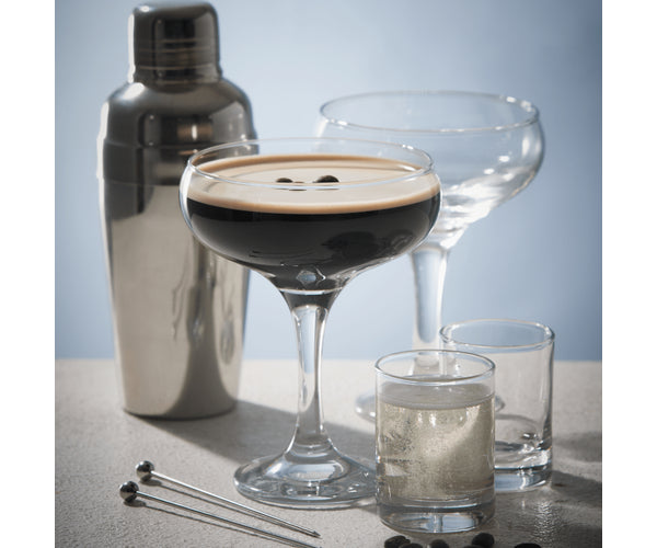 Two clear 20cl cocktail saucer glasses, one filled with a dark espresso martini, beside a metal shaker and two shot glasses.