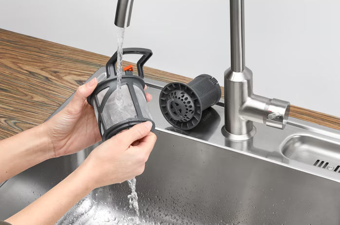 Hands rinsing a dishwasher filter under a running kitchen faucet next to a stainless steel sink and tap.